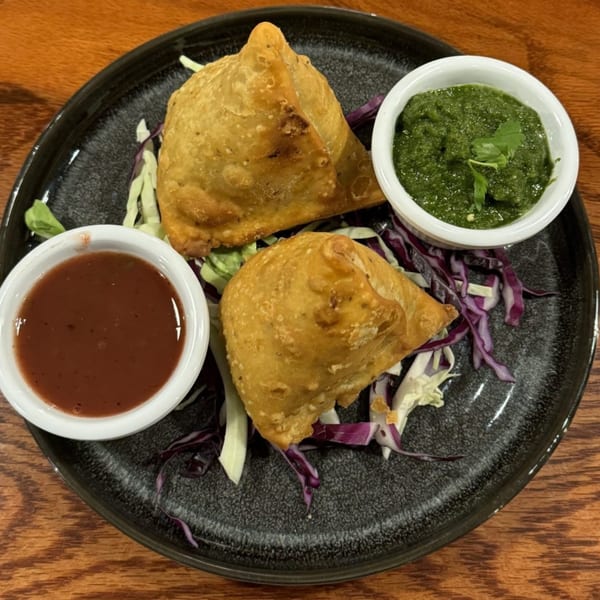 Two golden-brown samosas with green and red dipping sauces on a dark plate with shredded cabbage.
